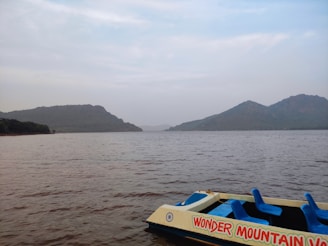 A serene lake with calm waters reflecting the overcast sky is flanked by rolling hills in the distance. A paddle boat with the words 'Wonder Mountain' is partially visible in the foreground, featuring bright blue seats.