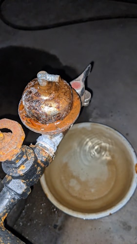 A rusted metal faucet with peeling surface, connected to a pipe. Below, a round container appears to be filled with liquid. The setting is dimly lit, suggesting an industrial or basement environment.