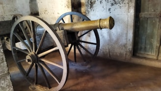 An old-fashioned cannon is mounted on large wooden wheels with intricate design work visible on the barrel. The cannon is situated indoors, against a textured stone wall with a wooden door partially visible on the side. The setting suggests a historical or museum environment.