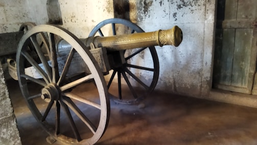 An old-fashioned cannon is mounted on large wooden wheels with intricate design work visible on the barrel. The cannon is situated indoors, against a textured stone wall with a wooden door partially visible on the side. The setting suggests a historical or museum environment.