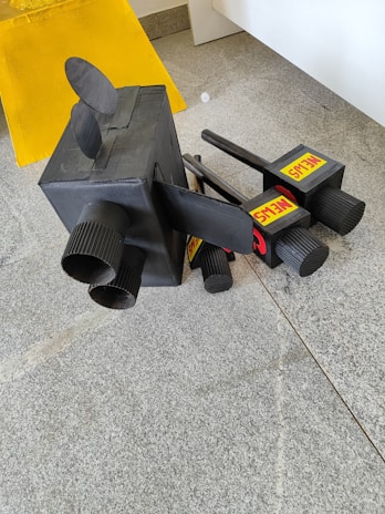 A collection of black camera-like objects placed on a gray tiled floor. The objects have various cylindrical protrusions and labels with text. A yellow board is situated in the background.