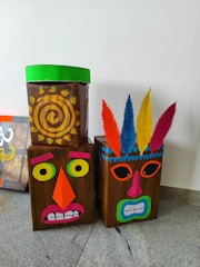 Three cardboard boxes are decorated as abstract, colorful faces. The first box has a spiral sun-like pattern with a green top. The second box features an orange triangular nose and exaggerated facial features. The third box includes vibrant feathers on top in various colors like blue, yellow, and red.