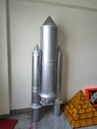 A model of a rocket made from metallic or aluminum-colored material stands upright. It has a central cylindrical body with two smaller cylindrical boosters attached to its sides. The rocket is positioned against a plain wall, and there is a pyramid-shaped structure with a brick-like pattern beside it.