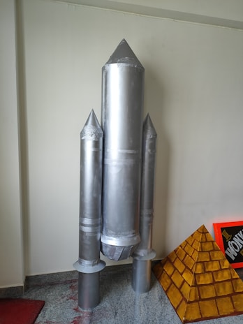 A model of a rocket made from metallic or aluminum-colored material stands upright. It has a central cylindrical body with two smaller cylindrical boosters attached to its sides. The rocket is positioned against a plain wall, and there is a pyramid-shaped structure with a brick-like pattern beside it.
