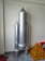 A model of a rocket made from metallic or aluminum-colored material stands upright. It has a central cylindrical body with two smaller cylindrical boosters attached to its sides. The rocket is positioned against a plain wall, and there is a pyramid-shaped structure with a brick-like pattern beside it.