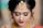 A woman adorned with traditional jewelry, featuring intricate gold designs on her forehead and a small nose ring. Her makeup is elegantly done with bold eyeliner, gold eyeshadow, and deep red lipstick. Her hair is styled in an elegant updo, complementing her overall appearance.