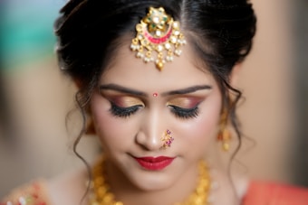 A woman adorned with traditional jewelry, featuring intricate gold designs on her forehead and a small nose ring. Her makeup is elegantly done with bold eyeliner, gold eyeshadow, and deep red lipstick. Her hair is styled in an elegant updo, complementing her overall appearance.