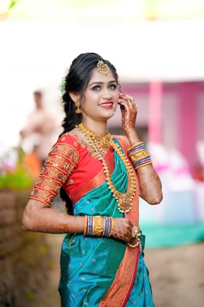 A woman wearing a traditional outfit stands gracefully, adorned with intricate jewelry. Her attire features a vibrant mix of turquoise and red with elaborate gold embroidery. Her hands display detailed henna designs, and her hair is styled elegantly with floral accents.