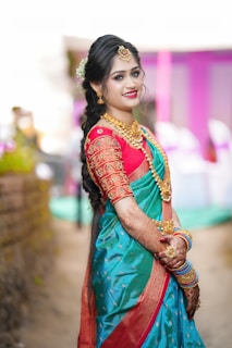 A woman dressed in vibrant traditional attire, including a colorful saree with intricate designs, stands smiling with her hands adorned with bangles and mehndi. She wears ornate jewelry including earrings, a necklace, and a maang tikka. Her hair is styled elegantly with floral embellishments.