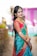 A woman dressed in vibrant traditional attire, including a colorful saree with intricate designs, stands smiling with her hands adorned with bangles and mehndi. She wears ornate jewelry including earrings, a necklace, and a maang tikka. Her hair is styled elegantly with floral embellishments.