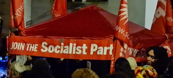 A red tent with the slogan 'JOIN THE Socialist Party' is surrounded by several red flags and people. The scene appears to be part of a public gathering or political rally, with multiple heads visible in the crowd.