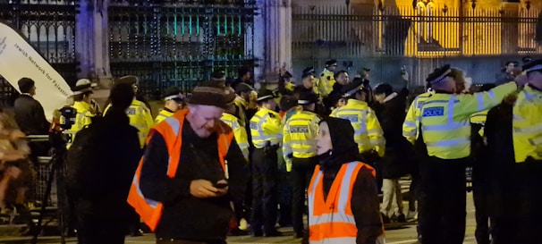 A group of police officers in high-visibility jackets stand in front of a large gated area, possibly a governmental or significant public building. In the foreground, a few individuals wearing orange safety vests are engaged in conversation. It appears to be an event or protest, with onlookers and participants gathered around.