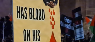 A protest scene with a prominent sign containing the words 'HAS BLOOD ON HIS' accompanied by an image of hands and red blood droplets. In the background, other protest signs such as 'Free Palestine' are partially visible.