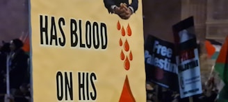 A protest scene with a prominent sign containing the words 'HAS BLOOD ON HIS' accompanied by an image of hands and red blood droplets. In the background, other protest signs such as 'Free Palestine' are partially visible.