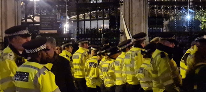 A line of uniformed police officers wearing bright yellow jackets stand in front of a tall metal gate. The scene is dimly lit, suggesting it is nighttime, and the officers seem to be attentive to something in front of them.