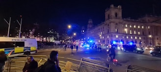 A bustling nighttime city scene with a large building illuminated in the background and several police vehicles with flashing blue lights. People are walking and biking on the street, and barriers are set up along the roadside. The atmosphere suggests a police presence for a public event or protest.