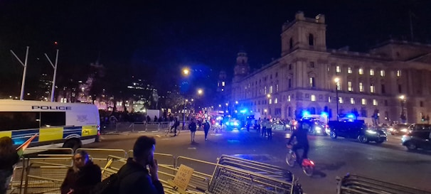 A bustling nighttime city scene with a large building illuminated in the background and several police vehicles with flashing blue lights. People are walking and biking on the street, and barriers are set up along the roadside. The atmosphere suggests a police presence for a public event or protest.
