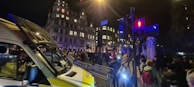 A nighttime urban scene involving a crowd of people near a large, illuminated building. A police van is parked on the street, and metal barricades are set up alongside. Traffic lights are visible, casting red and blue glows. The atmosphere is busy, with pedestrians walking past.