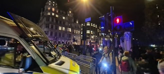 A nighttime urban scene involving a crowd of people near a large, illuminated building. A police van is parked on the street, and metal barricades are set up alongside. Traffic lights are visible, casting red and blue glows. The atmosphere is busy, with pedestrians walking past.