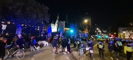 A nighttime street scene features a large group of people, some on bicycles, and several police officers wearing reflective vests. There are streetlights illuminating the area, and historic buildings in the background. Some individuals appear to be holding signs or banners, suggesting a protest or public gathering.