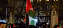 A group of people is gathered outside at night, holding flags with red, white, and green colors. The scene is set in front of a dark building with decorative architecture, and there are iron fences in the foreground. The lighting is dim, and the image has a slightly subdued atmosphere.