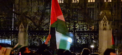 A group of people is gathered outside at night, holding flags with red, white, and green colors. The scene is set in front of a dark building with decorative architecture, and there are iron fences in the foreground. The lighting is dim, and the image has a slightly subdued atmosphere.