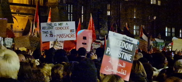 A large crowd gathered at night, holding various protest signs and flags. The signs convey messages related to political issues, such as 'Freedom for Palestine' and statements about genocide. The environment is dimly lit, likely illuminated by streetlights.