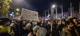 A large crowd of people is gathered for a protest at night. Many individuals are holding signs, one prominently displaying a message about human suffering and dehumanization. The scene is illuminated by streetlights, and several flags are visible in the background.