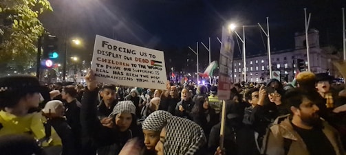A large crowd of people is gathered for a protest at night. Many individuals are holding signs, one prominently displaying a message about human suffering and dehumanization. The scene is illuminated by streetlights, and several flags are visible in the background.