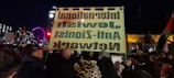 A night scene captures a protest or demonstration with a large crowd gathered. People are holding signs and flags, and a significant banner reads 'International Jewish Anti-Zionist Network' in reverse. The background features notable landmarks such as a brightly lit Ferris wheel and a clock tower, likely indicating a central urban location.