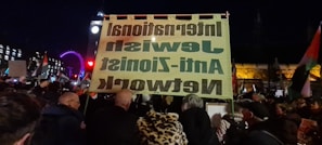 A night scene captures a protest or demonstration with a large crowd gathered. People are holding signs and flags, and a significant banner reads 'International Jewish Anti-Zionist Network' in reverse. The background features notable landmarks such as a brightly lit Ferris wheel and a clock tower, likely indicating a central urban location.
