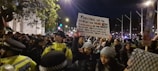 A large group of people, some wearing black-and-white keffiyeh headscarves, are gathered in a nighttime protest. One woman prominently holds a sign with a message about displaying dead children to prove suffering. Police officers in yellow jackets are present, managing the crowd. The scene is illuminated by streetlights and city lights, indicating an urban environment.