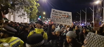 A large group of people, some wearing black-and-white keffiyeh headscarves, are gathered in a nighttime protest. One woman prominently holds a sign with a message about displaying dead children to prove suffering. Police officers in yellow jackets are present, managing the crowd. The scene is illuminated by streetlights and city lights, indicating an urban environment.