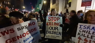 A street protest with people holding signs and banners advocating against violence towards children. The crowd is visibly engaged, with various individuals holding placards with strong messages. The setting appears to be an urban area at night, with dim lighting and a sense of urgency and determination among the participants.