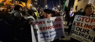 A crowd of people is gathered, holding protest signs. The signs have messages about genocide and stopping violence against children, related to Palestine. The scene is set at night, possibly on a busy street or outside of a building.