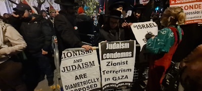 A group of people are gathered at a protest, holding signs with messages related to Zionism and Judaism. The signs read slogans such as 'Zionism and Judaism are diametrically opposed' and 'Judaism condemns Zionist terrorism in Gaza.' The crowd appears to be diverse, with individuals dressed warmly, some wearing hats. The surrounding environment suggests it is taking place outdoors, perhaps on a city street at night, indicated by the dim lighting and presence of trees in the background.