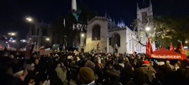 A large crowd of people gathered at night in front of a historic building, with many individuals holding flags and placards. The area is illuminated by streetlights, and a red tent displays the words 'Socialist Party'. Trees are visible alongside the building.