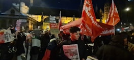 A nighttime protest scene with a large crowd holding signs and banners. Prominent red flags display the word 'Socialist,' and various signs convey messages, including 'Freedom for Palestine.' The background features an illuminated historic building, suggesting a significant location for the protest.