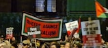 A crowd of people holding various protest signs, including 'Queers Against Imperialism,' 'Free Palestine,' and 'Stop the War on Gaza,' during a nighttime demonstration. The image captures the energy and activism of the protest with multiple signs being raised in a busy and dynamic scene.