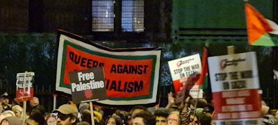 A crowd of people holding various protest signs, including 'Queers Against Imperialism,' 'Free Palestine,' and 'Stop the War on Gaza,' during a nighttime demonstration. The image captures the energy and activism of the protest with multiple signs being raised in a busy and dynamic scene.