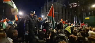 A large group of people are gathered outside at night, holding flags with red, black, white, and green colors. The crowd seems actively engaged, some people are on elevated positions, and there is a building in the background lit up but largely in shadows.