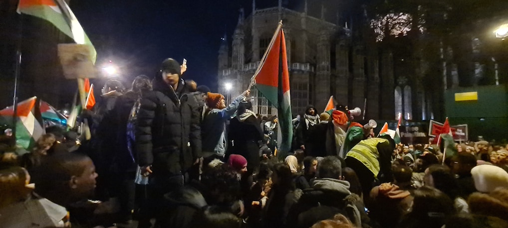 A large group of people are gathered outside at night, holding flags with red, black, white, and green colors. The crowd seems actively engaged, some people are on elevated positions, and there is a building in the background lit up but largely in shadows.