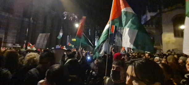 A large gathering of people in a nighttime outdoor setting, holding and waving large flags with red, white, black, and green colors. The crowd appears dense and is illuminated by streetlights. Several people are facing in different directions, and some are partially obscured by others.