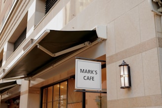 A building facade with a prominent sign reading 'MARK'S CAFE'. The exterior features a light stone material with a black awning above the windows. An outdoor light fixture is mounted on the wall next to the sign.