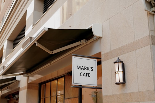 A building facade with a prominent sign reading 'MARK'S CAFE'. The exterior features a light stone material with a black awning above the windows. An outdoor light fixture is mounted on the wall next to the sign.