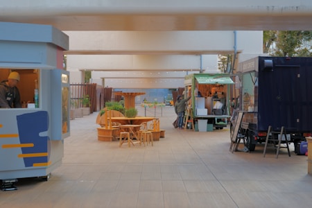 A small open-air market or food court is situated under a modern overpass. Several food stalls and trucks line either side of a wide pedestrian walkway. A few wooden tables and chairs are centrally positioned. Two individuals are visible at different stalls, engaged in setting up or serving.