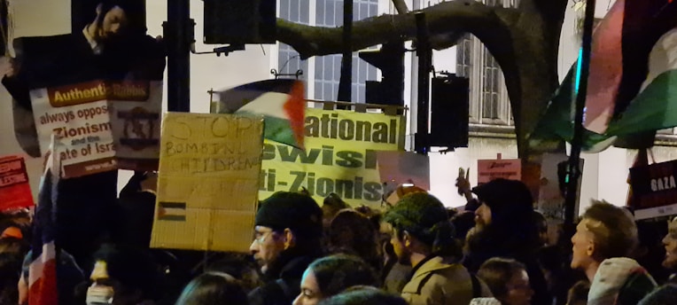 A nighttime protest with a crowd holding signs and flags. The focus is on several placards with messages about anti-Zionism and children's rights. People in the crowd are densely packed, some holding up banners and wearing hats or hoodies.
