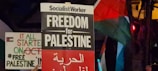 Protest signs advocating for Palestine are visible, with one prominently displaying 'Freedom for Palestine' along with Arabic text. A flag can be seen in the background, likely the Palestinian flag with red, green, and black colors.
