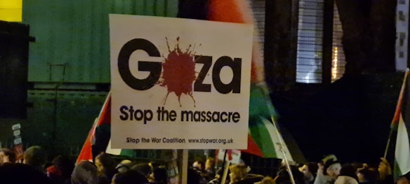 A protest scene with a prominent sign in the foreground reading 'Gaza Stop the massacre' accompanied by a blood splatter graphic. The sign includes the logo of the Stop the War Coalition. Background elements include a crowd and some flags, partially visible and waving, suggesting an outdoor setting.