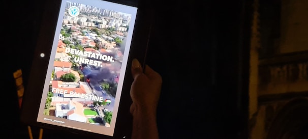A person holds a tablet displaying an aerial view of a city with visible smoke and destruction. The screen includes text saying 'Devastation. Unrest. Free Palestine.' along with an emblem and hashtags.
